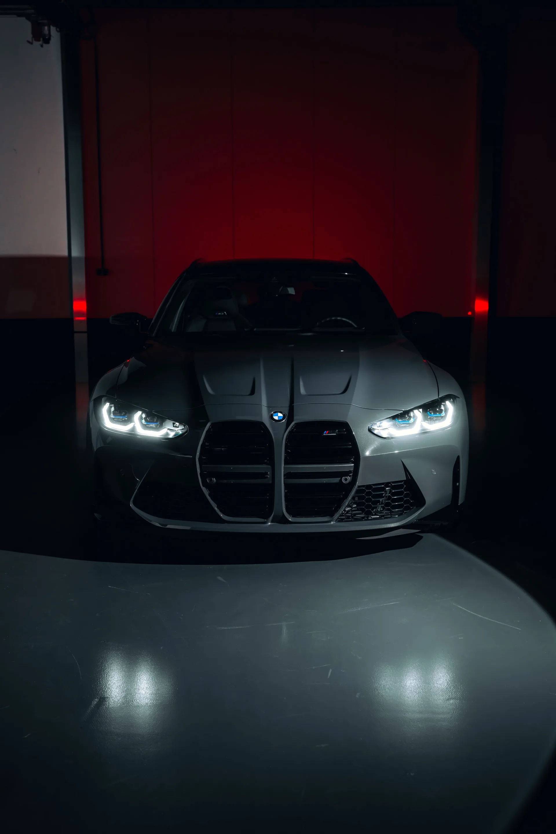 Grijze BMW M3 Touring recht van voren in een donkere studio met rood licht